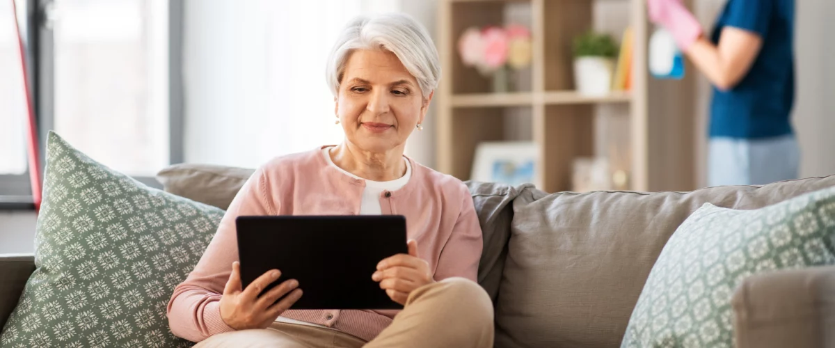 femme âgée heureuse avec une tablette et une aide à domicile qui nettoie sa maison - menageparfait.fr